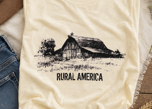 A cream-colored t-shirt with a black graphic of a barn and the text 'RURAL AMERICA', styled in a flat-lay with jeans, a hat, and dried wheat on a wooden surface.