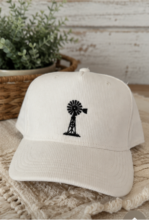 Two corduroy baseball caps shown side by side in a split-screen layout. The left cap is medium blue with a black embroidered floral design, and the right cap is white with a black embroidered windmill design.