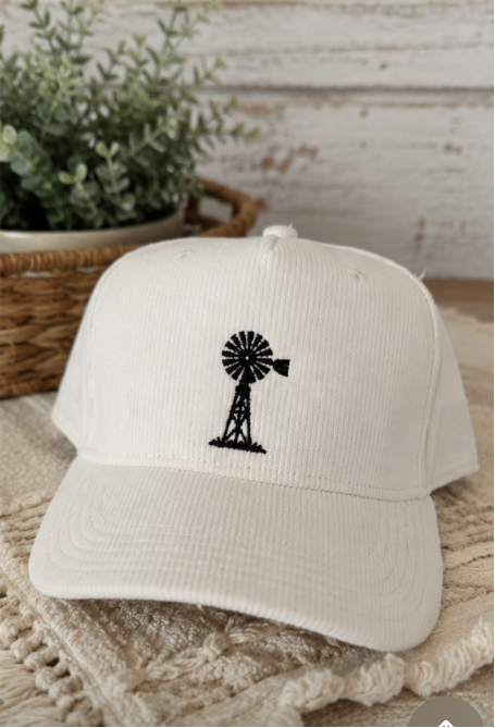 Two corduroy baseball caps shown side by side in a split-screen layout. The left cap is medium blue with a black embroidered floral design, and the right cap is white with a black embroidered windmill design.